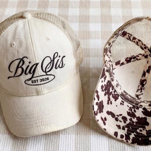 Puede incluir: Dos gorras de béisbol. Una es de color crema con la parte trasera de malla y las palabras "Big Sis EST 2026" en escritura negra. La otra gorra tiene un diseño de estampado de vaca marrón y blanco en el interior y la parte trasera de malla.