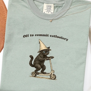 Op de afbeelding: Lichtgroen t-shirt met de tekst "Off to commit catfoolery" en een afbeelding van een zwarte kat met een tovenaars hoed op een scooter. Het shirt heeft een ronde hals en korte mouwen.
