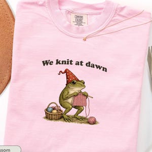 May include: A light pink t-shirt with the text "We knit at dawn" above an illustration of a frog knitting. The frog wears a red and white polka-dot hat and sits next to a basket of yarn. The shirt has a Comfort Colors tag.