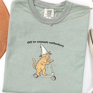 Op de afbeelding: Lichtgroen t-shirt met de tekst "Off to commit catfoolery" en een cartoonkat met een tovenaars hoed op een scooter. Het shirt heeft een ronde hals en korte mouwen. Een gouden ketting met een ster hanger ligt op het shirt.