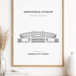May include: Framed black and white print of Arrowhead Stadium, home of the Kansas City Chiefs. The print includes the stadium's name, coordinates, and team name. The artwork is displayed on a wooden cabinet with decorative items.