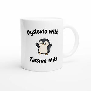 Op de afbeelding: Witte keramische mok met een cartoon pinguïn. De mok heeft de tekst "Dyslexic with Tassive Mits" in zwarte letters. De pinguïn is zwart en wit met gele voeten.