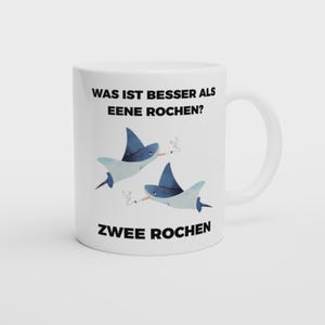 Könnte beinhalten: Weiße Keramik-Tasse mit dem Text "WAS IST BESSER ALS EENE ROCHEN? ZWEE ROCHEN". Die Tasse zeigt eine Cartoon-Illustration von zwei blauen Rochen, die Zigaretten rauchen.