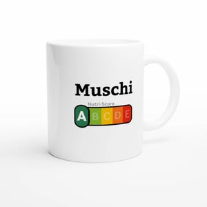 Könnte beinhalten: Eine weiße Keramiktasse mit dem schwarzen Schriftzug Muschi über einer Nutri-Score-Grafik. Die Grafik zeigt einen horizontalen Balken mit den Buchstaben A, B, C, D und E in Grün, Hellgrün, Gelb, Orange und Rot, wobei das A grün hervorgehoben ist.
