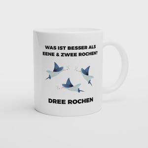 Könnte beinhalten: Weiße Keramik-Tasse mit einem humorvollen Design. Die Tasse trägt den Text "WAS IST BESSER ALS EENE & ZWEE ROCHEN?" und "DREE ROCHEN" mit Illustrationen von drei blauen Rochen. Die Tasse hat einen weißen Henkel.