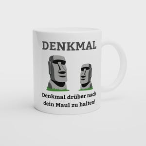 Könnte beinhalten: Ein weißer Keramikbecher mit einer Grafik von zwei grauen Moai-Steinstatuen auf kleinen grünen Grasflächen. Der Text DENKMAL ist in Grau über den Statuen aufgedruckt, und der Text Denkmal drüber nach dein Maul zu halten! steht darunter.