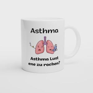 Peut inclure: Un mug en céramique blanc avec une illustration de deux poumons roses. Un poumon tient une cigarette allumée, l'autre un inhalateur bleu. Le texte indique Asthma en haut et Asthma Lust ene zu rochen? en bas du motif central.