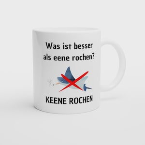 Könnte beinhalten: Weiße Keramik-Tasse mit einem humorvollen Design. Die Tasse zeigt einen blauen Cartoon-Rochen mit einer durchgestrichenen Zigarette. Der Text auf Deutsch lautet "Was ist besser als eene rochen?" und "KEENE ROCHEN."