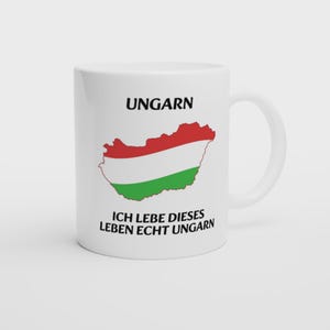 Könnte beinhalten: Weiße Keramik-Tasse mit dem Wort "UNGARN" und einer Karte von Ungarn in Rot, Weiß und Grün. Der Text "ICH LEBE DIESES LEBEN ECHT UNGARN" steht darunter. Die Tasse hat einen gebogenen Griff.