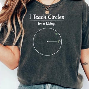 Puede incluir: Camiseta gris oscuro con el texto "I Teach Circles for a Living". Un círculo blanco con un radio etiquetado como "r" y el símbolo pi está impreso en la camiseta. La camiseta es de estilo casual.