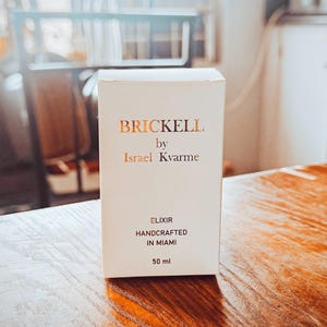 Op de afbeelding: Een witte productdoos met gouden letters voor "BRICKELL by Israel Kvarme" wordt gepresenteerd op een houten oppervlak. De doos vermeldt ook "ELIXIR HANDCRAFTED IN MIAMI" en "50 ml". De achtergrond is wazig.