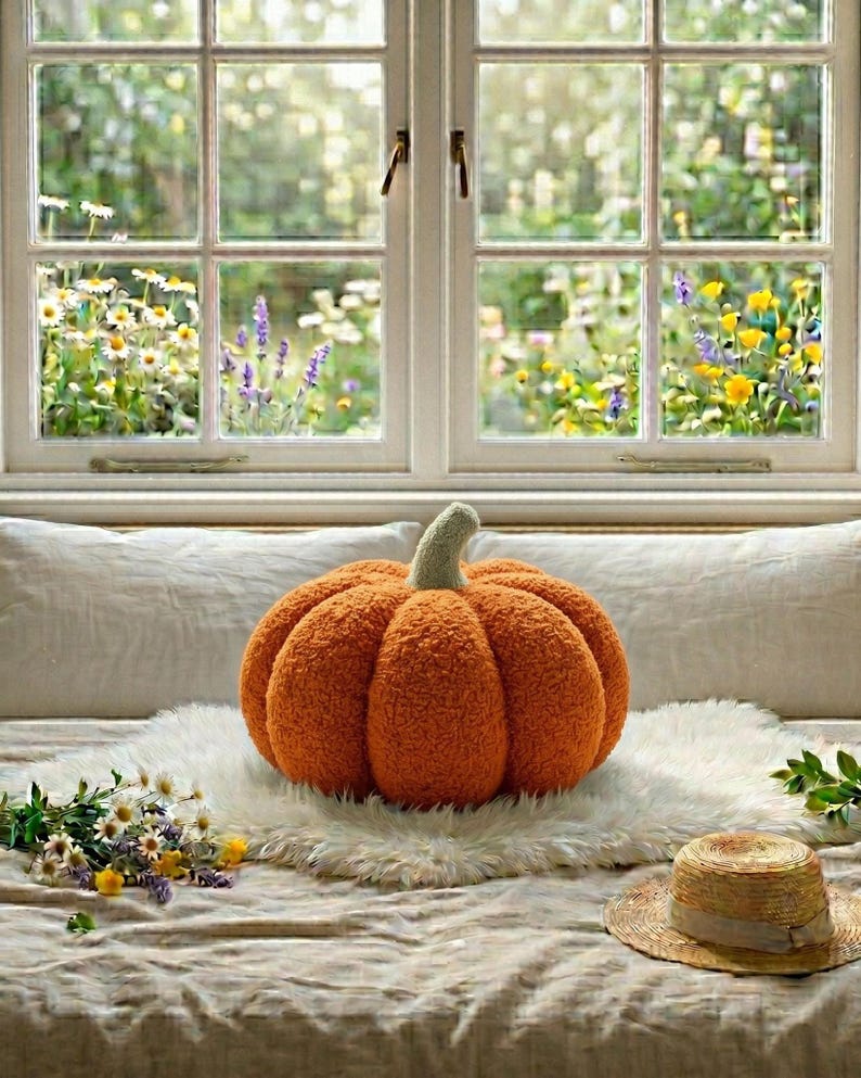 Pode incluir: Uma almofada redonda, laranja e texturizada em forma de ab&oacute;bora com um caule verde-claro. A almofada repousa sobre um tapete de pele sint&eacute;tica branca, acompanhada por um chap&eacute;u de palha e flores silvestres numa superf&iacute;cie de tecido neutro.