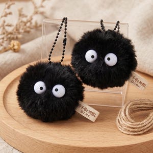 Fluffy soot sprite plushie keychain resting on a rustic wooden slice, surrounded by linen fabric and crafting tools, highlighting the artisan handmade quality