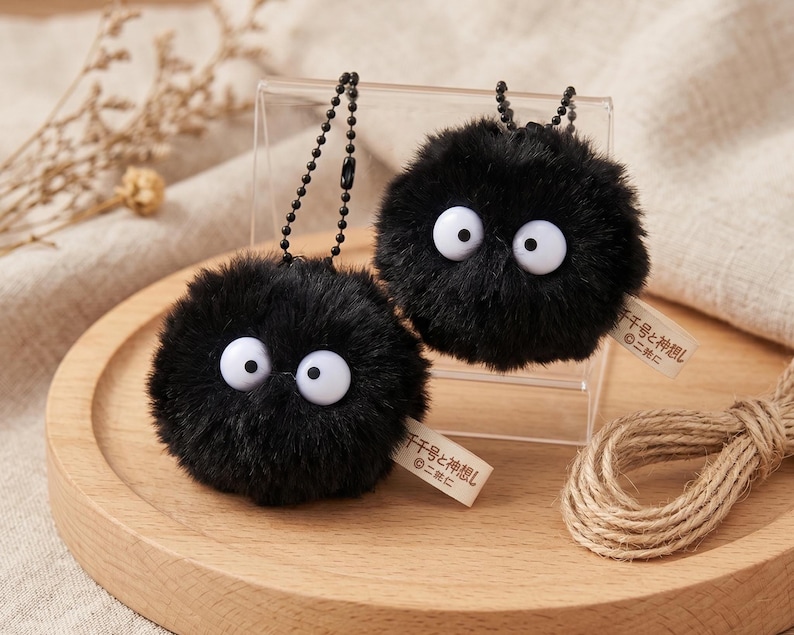 Fluffy soot sprite plushie keychain resting on a rustic wooden slice, surrounded by linen fabric and crafting tools, highlighting the artisan handmade quality