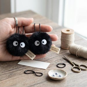 Detailed close-up of a handmade black dust spirit keychain with large white googly eyes and a soft faux-fur texture, showing the high-quality craftsmanship