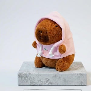 Peut inclure: Une peluche capybara marron portant un sweat à capuche rose clair avec un motif de pêche sur le devant et des cordons blancs. Le texte ci-dessous indique : COZY CAPYBARA PEACH HOODIE PLUSH | HANDCRAFTED | ETSY EXCLUSIVE. La peluche est douce.