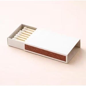 May include: A white matchbox with a brown striking surface and a partially open lid. The box contains several matches with white tips. The background is a solid, light pink color.