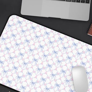 May include: A rectangular desk mat featuring a repeating pattern of light blue bows and small pink flowers on a white background. The mat has a black border and is positioned on a dark surface with a laptop, a mouse, a notebook, and a coffee mug.
