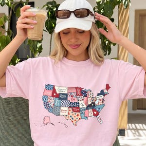 May include: A light pink short-sleeve t-shirt featuring a graphic of the United States map filled with a patchwork of red, white, and blue patterns, including stars, stripes, and floral designs, with the word Blossom printed in black text.