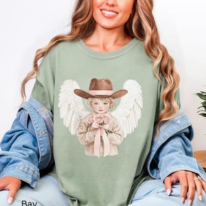 May include: Sage green t-shirt featuring a graphic of a child angel wearing a brown cowboy hat and holding pink roses. The angel has white wings and is tied with a pink ribbon. The word "Bay" is printed at the bottom.