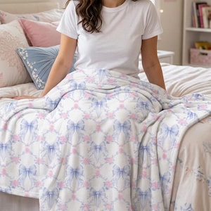 May include: A soft white fleece blanket featuring a repeating pattern of light blue bows and small pink floral clusters. The fabric has a plush texture and is draped over a bed, showcasing the symmetrical design of the bows and flowers throughout.
