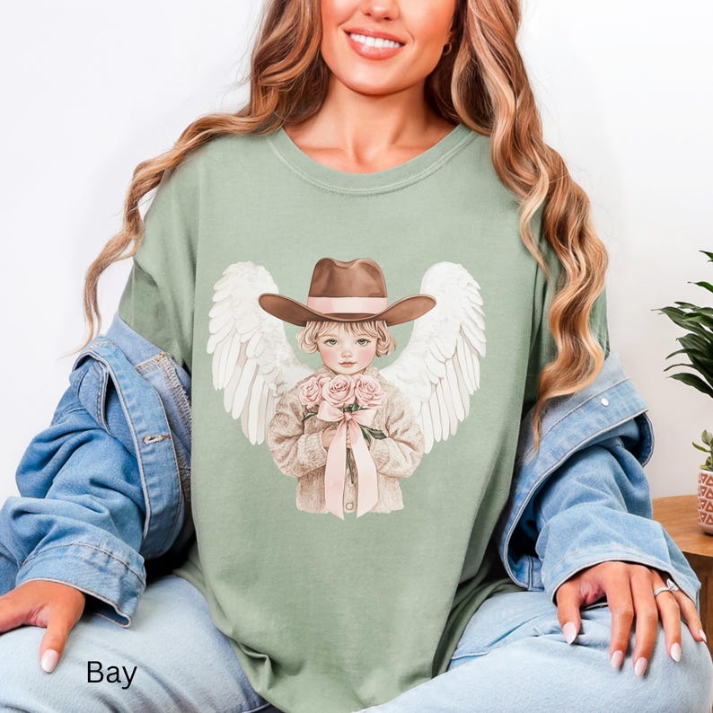May include: Sage green t-shirt featuring a graphic of a child angel wearing a brown cowboy hat and holding pink roses. The angel has white wings and is tied with a pink ribbon. The word "Bay" is printed at the bottom.