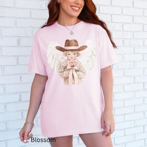 May include: A pale pink t-shirt with a graphic of a child angel in a cowboy hat, holding roses and with white wings. The word "Blossom" is printed at the bottom of the shirt.
