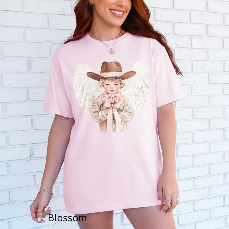 May include: A pale pink t-shirt with a graphic of a child angel in a cowboy hat, holding roses and with white wings. The word "Blossom" is printed at the bottom of the shirt.