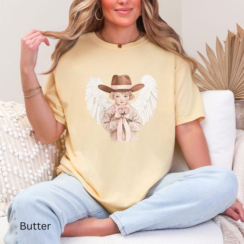 May include: Pale yellow t-shirt with a watercolour illustration of a child angel wearing a cowboy hat and holding roses. The angel has white wings. The word "Butter" is printed at the bottom of the shirt.