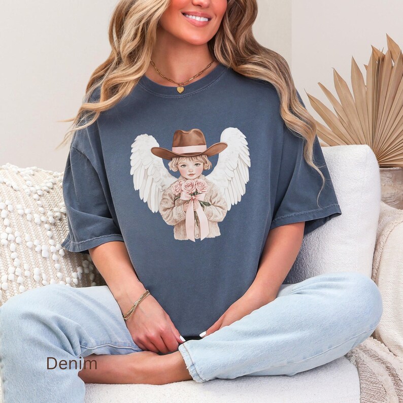 May include: Denim blue t-shirt with a graphic of an angel wearing a cowboy hat and holding roses. The angel has white wings. The word "Denim" is printed at the bottom of the shirt.