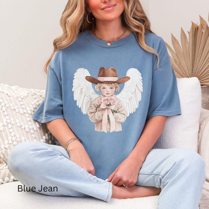 May include: A light blue t-shirt with a graphic of an angel in a cowboy hat, holding roses and with white wings. The text "Blue Jean" is at the bottom. The shirt is a relaxed fit, casual style, suitable for everyday wear.