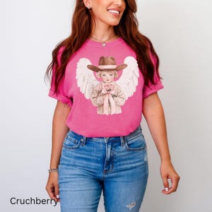 May include: A cerise t-shirt with a graphic of an angel in a cowboy hat, holding roses, and with white wings. The text "Cruchberry" is printed at the bottom of the shirt.