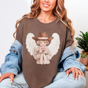 May include: Espresso-coloured t-shirt with a graphic of an angel wearing a cowboy hat and holding pink roses. The angel has white wings and is centered on the shirt. The word "Espresso" is printed at the bottom of the shirt.