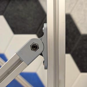 May include: Close-up of a silver-colored metal hinge connecting two aluminum bars. The hinge is secured with a bolt and is attached to a vertical aluminum bar. The background features a geometric tile pattern in shades of gray, white, and blue.