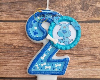 Könnte beinhalten: Blue's Clues Geburtstagskerze in Form der Zahl zwei. Die Kerze ist weiß mit blauem und weißem Glitzer und hat ein blaues Pfotenabdruck-Design. Die Kerze hat ein blaues und weißes Bild von Blue dem Hund oben drauf.