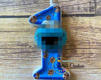 May include: Blue and white birthday candle shaped like the number one. The candle is decorated with cookie images and the text "Kathy's Creations".