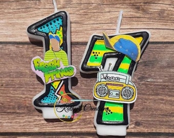 May include: Two birthday candles, one shaped like the number 1 and the other shaped like the number 7. The number 1 candle is decorated with a cartoon image of a person wearing a hat and the words "Fresh Prince". The number 7 candle is decorated with a cartoon image of a boombox and a hat.