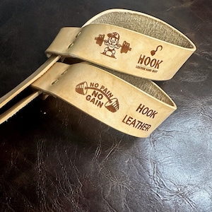 Puede incluir: Un par de ganchos de cuero de color beige claro para levantamiento de pesas con bordes cosidos. Los ganchos presentan el texto "HOOK LEATHER HAND CRAFT" y "NO PAIN NO GAIN". También se ve un gráfico de un levantador de pesas de dibujos animados.
