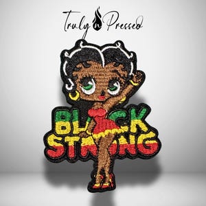May include: An embroidered patch featuring a cartoon character with black hair, green eyes, and a red dress, positioned over the text "BLACK STRONG" in green, yellow, and red. The brand name "Truly Pressed" appears in black script above the patch.