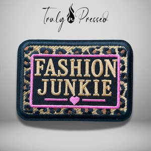 May include: A rectangular embroidered patch featuring a black background with a gold and black leopard print border. The center displays the text FASHION JUNKIE in gold letters, enclosed by a pink rectangular border and a small pink heart icon.