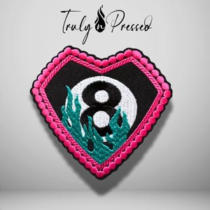 May include: A heart-shaped embroidered patch featuring a black background with a white eight ball graphic surrounded by teal flames. The patch is bordered by a bright pink scalloped edge. The text Truly Pressed is visible above the patch.