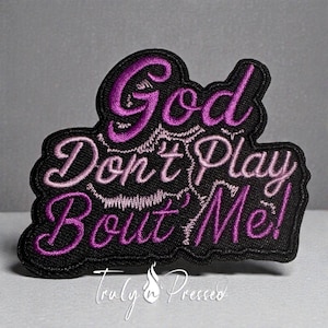 May include: A black embroidered patch with the phrase "God Don't Play Bout' Me!" in purple and pink thread. The patch has a decorative border and the text "Truly n Pressed" at the bottom.
