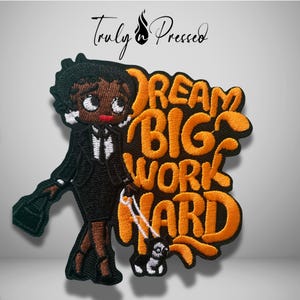 May include: An embroidered patch featuring a cartoon character in a black suit and skirt, holding a black bag and a white leash attached to a small dog. The text DREAM BIG WORK HARD is stitched in bold orange letters next to the character.
