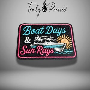 May include: A rectangular embroidered patch with a black background and a pink border. It features the text 'Boat Days & Sun Rays' in light blue and pink, along with an embroidered white boat, a yellow sun, and light blue water waves.