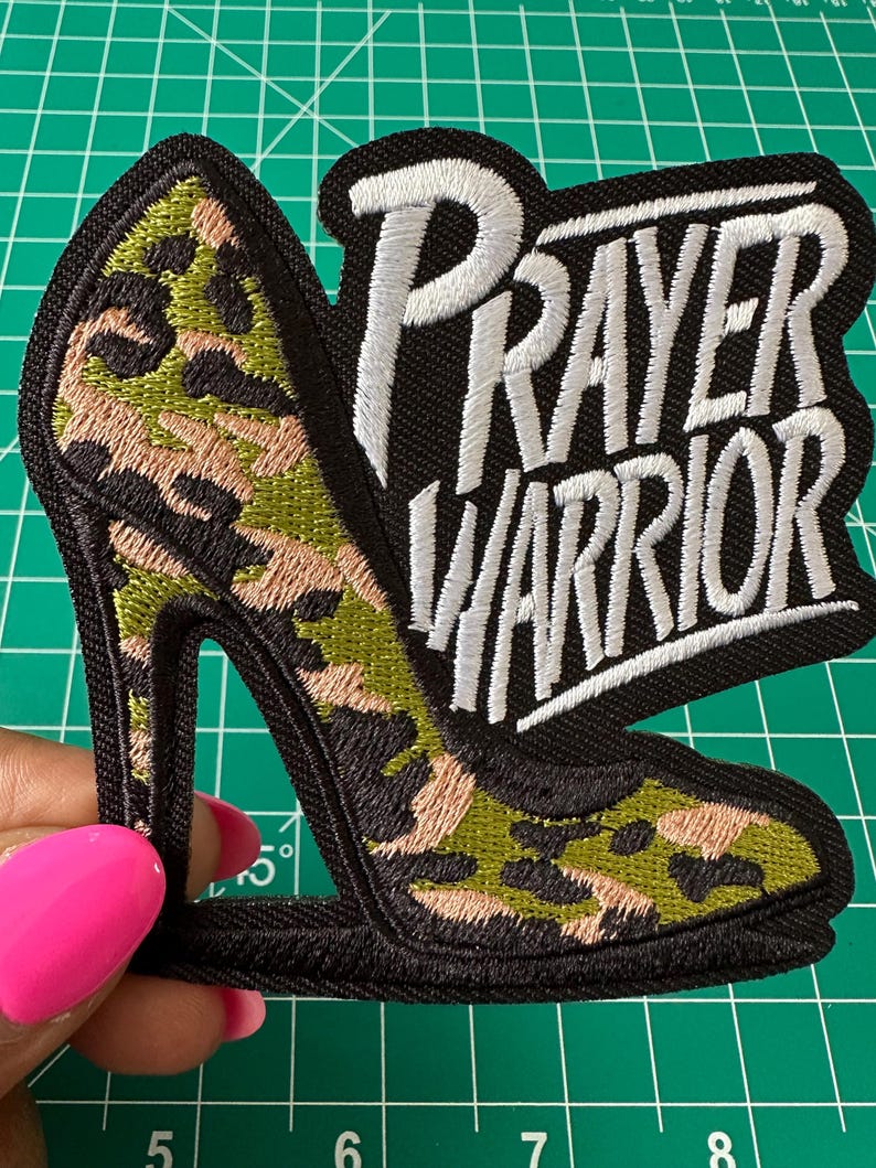 May include: An embroidered patch featuring a high-heeled shoe with a green, tan, and black camouflage pattern. The text "PRAYER WARRIOR" is stitched in white thread with a black background. The patch has a thick black border and measures over 3 inches.