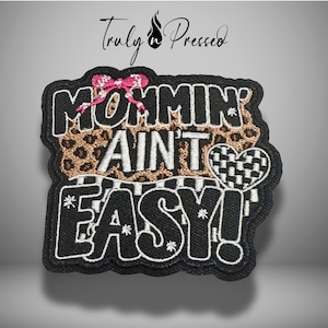 May include: Embroidered patch with the phrase "Mommin' Ain't Easy!" in white lettering on a black background. Features a pink bow, leopard print, and a checkered heart design. The patch is edged with a black border.