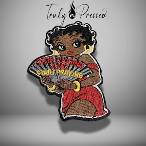 May include: An embroidered patch featuring a cartoon character with black hair, wearing a red dress and gold hoop earrings, holding a black fan with the red and yellow text "STOP JUDGING AND START PRAYING" written across the surface of the fan.