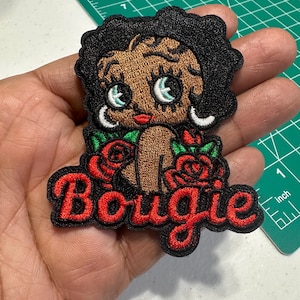 May include: An embroidered patch featuring a cartoon character with dark skin, black hair, and large eyes, surrounded by red roses with green leaves. The word Bougie is stitched in red cursive lettering at the bottom against a black background.