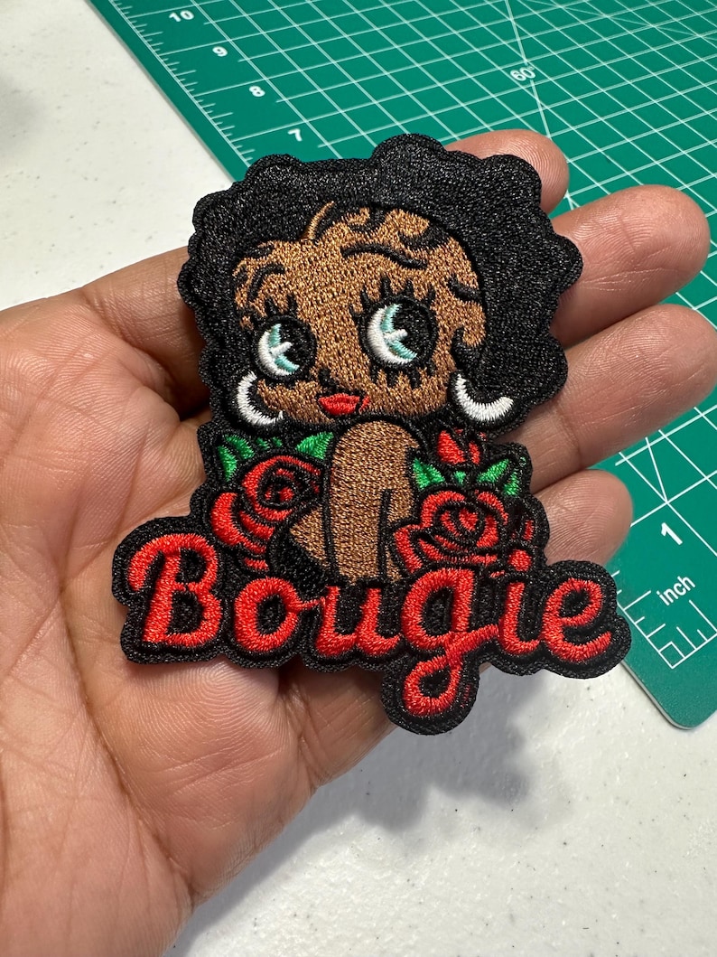May include: An embroidered patch featuring a cartoon character with dark skin, black hair, and large eyes, surrounded by red roses with green leaves. The word Bougie is stitched in red cursive lettering at the bottom against a black background.