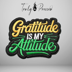 May include: An embroidered patch with a black background featuring the yellow text Gratitude, the white text IS MY, and the green text Attitude. The design includes decorative green lines and the text Truly Pressed at the top of the patch.
